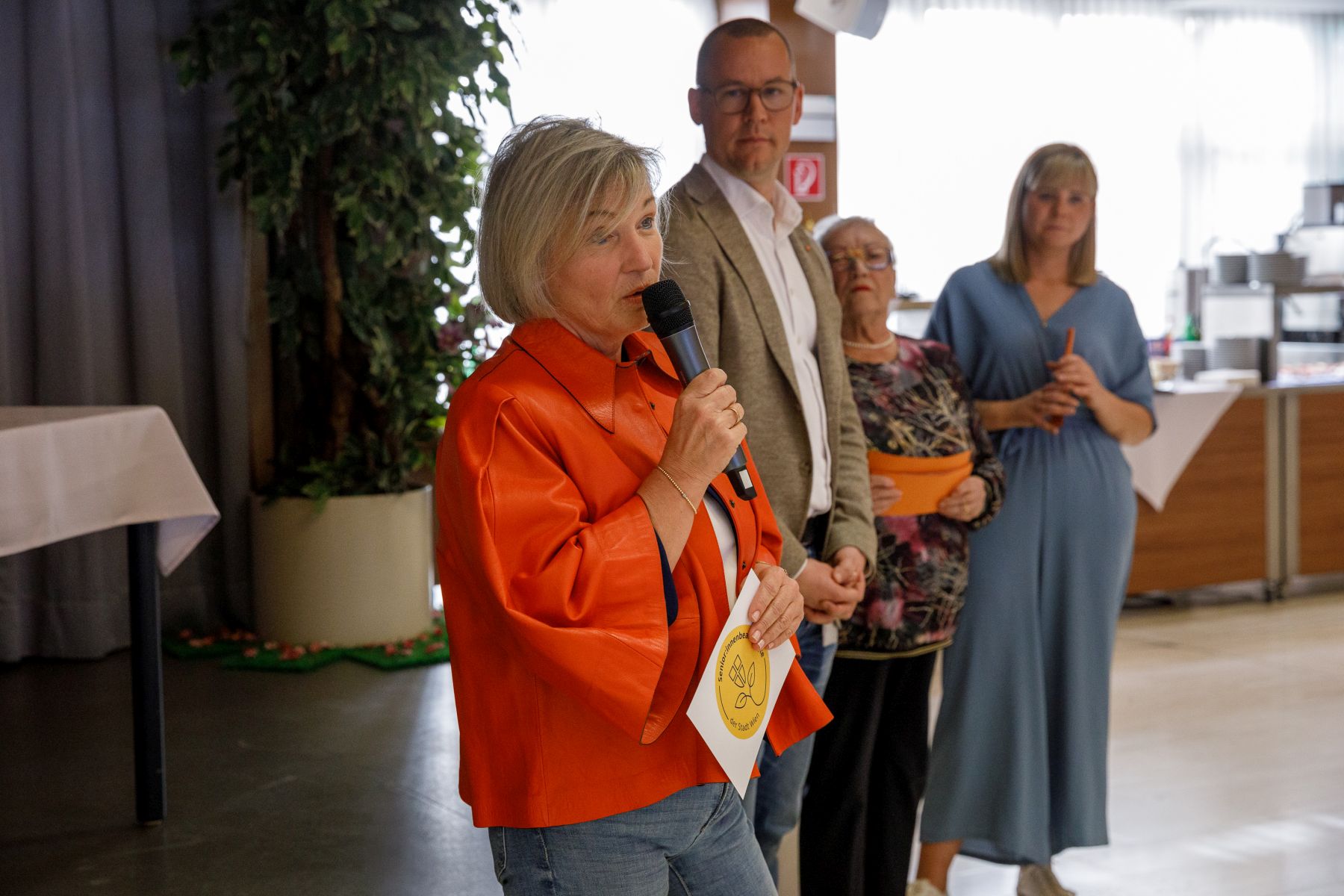 Sabine Hofer-Gruber, Senior*innen-Beauftragte für die Stadt Wien, hält eine Ansprache vor der Bühne, dahinter stehen Simon Bluma, stellvertretender Geschäftsführer Häuser zum Leben und die beiden Moderatorinnen der Preisverleihung