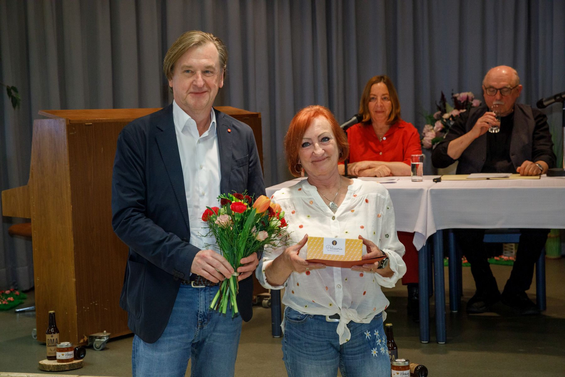 Christian Hennefeind, Geschäftsführer Häuser zum Leben mit einer Preisträgerin des Literatur-Contests 2026. Im Hintergrund sitzen die Schauspieler*innen Susi Stach und Karl Fischer.