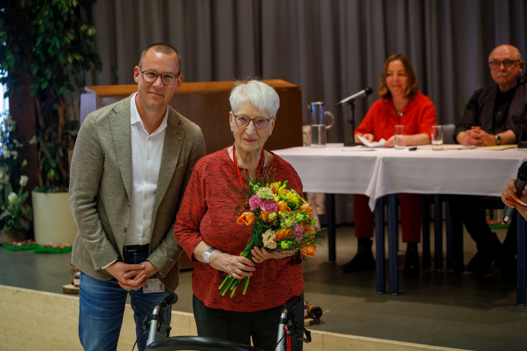 Simon Bluma, stellvertretender Geschäftsführer Häuser zum Leben mit Preisträgerin Traude Veran. Im Hintergrund sitzen die Schauspieler*innen Susi Stach und Karl Fischer.