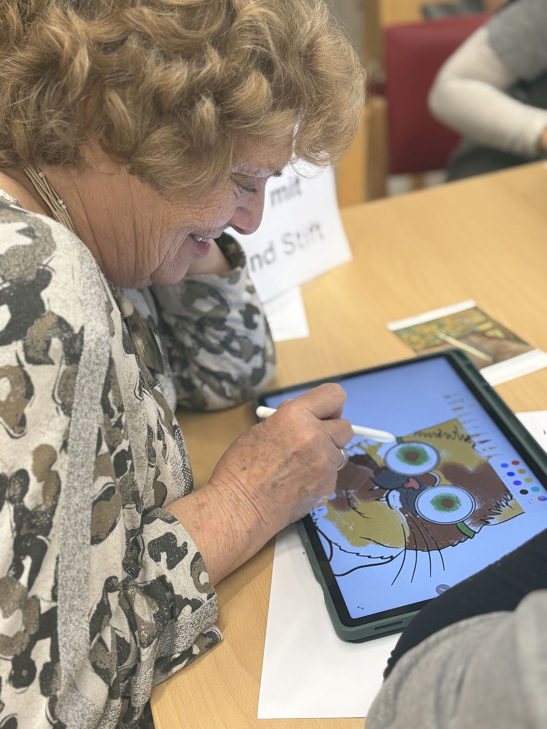 Eine Seniorin zeichnet eine Katze mit Brille auf einem Tablet-PC und lächelt.