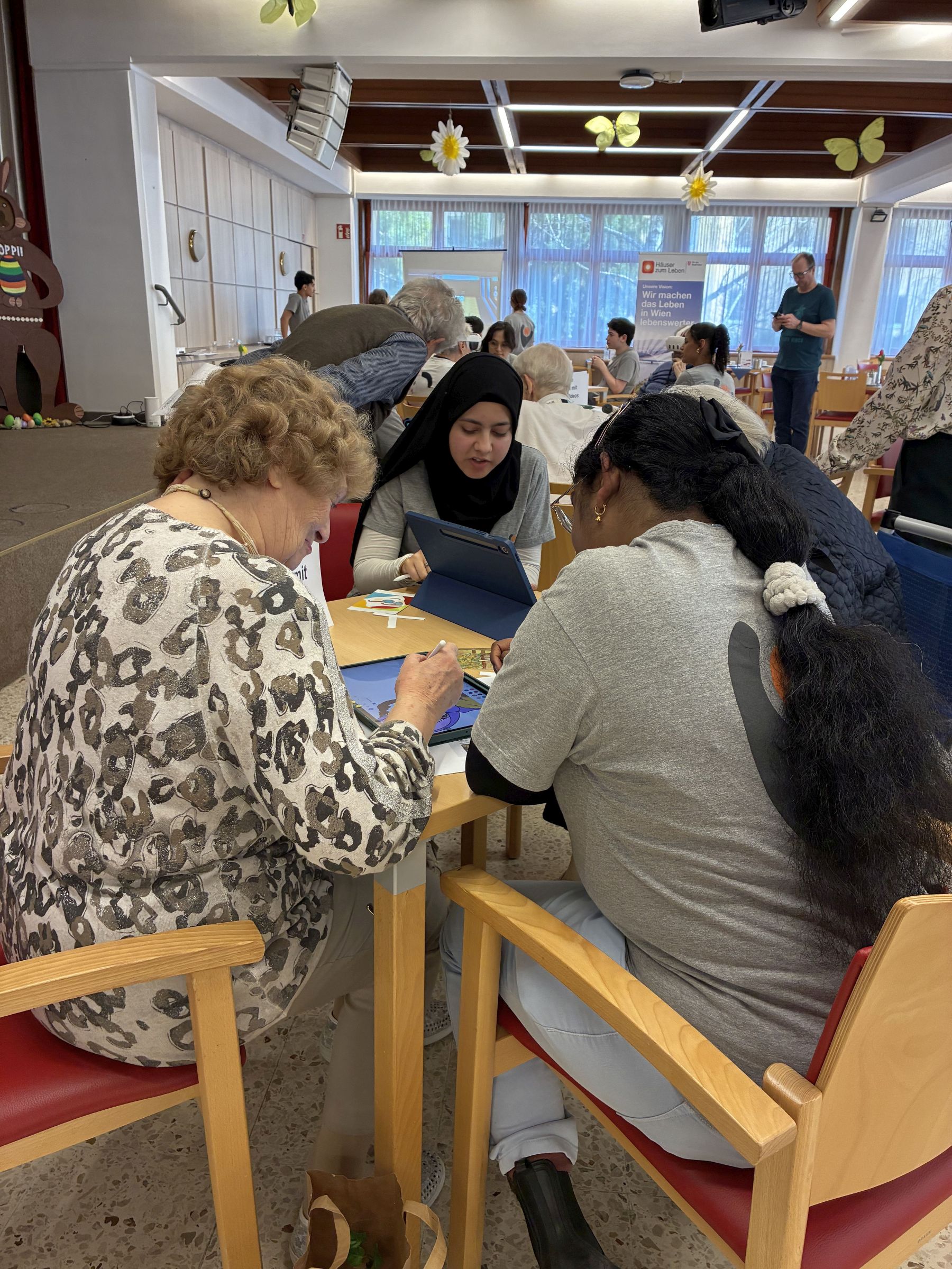 Jugendliche sitzen mit einer Seniorin am Tisch, die etwas auf einem Tablet-PC macht.