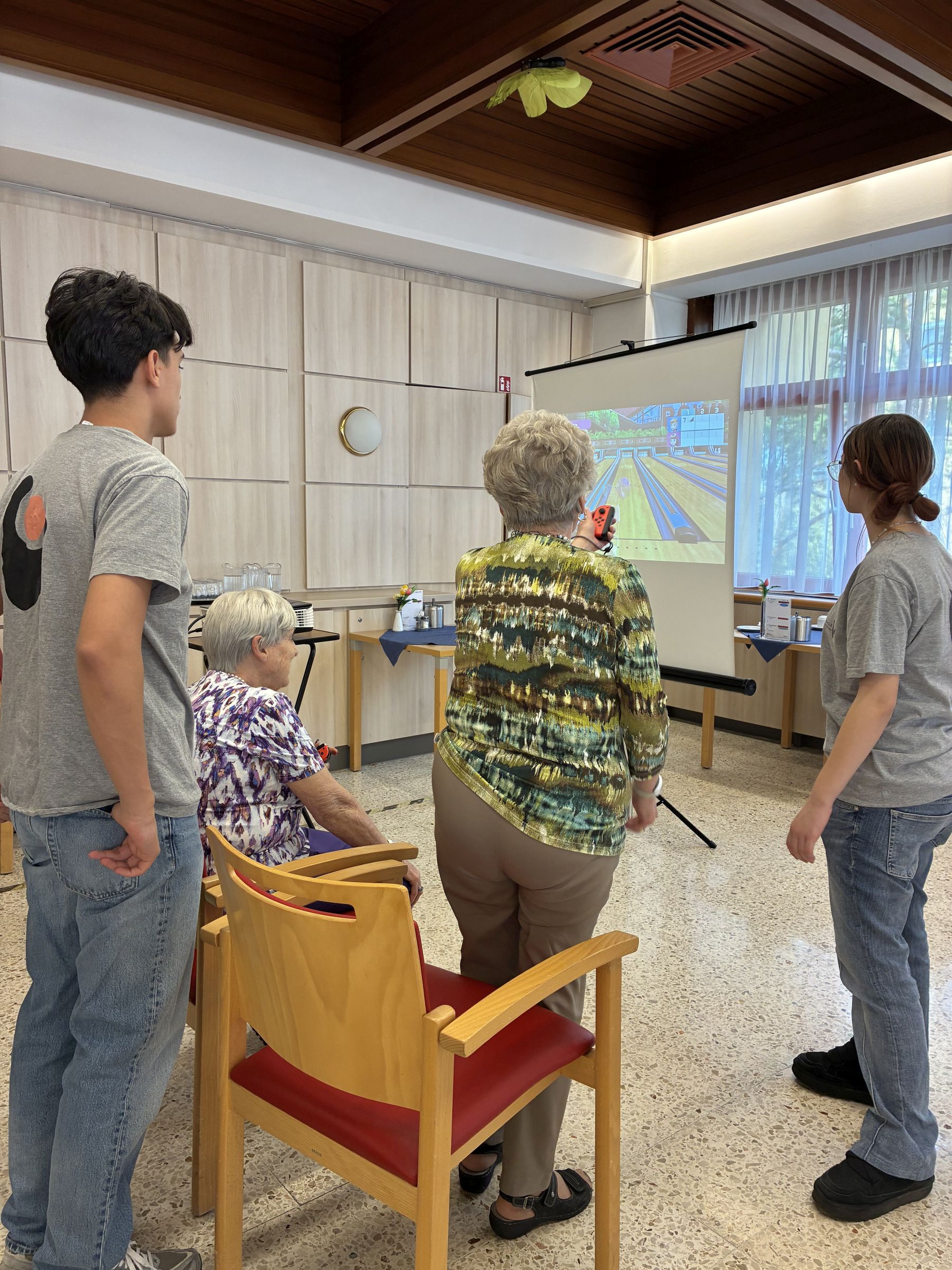 Zwei Jugendliche und zwei Seniorinnen von hinten, eine Seniorin hält einen Joystick und spielt ein Spiel, das auf einer Leinwand zu sehen ist.
