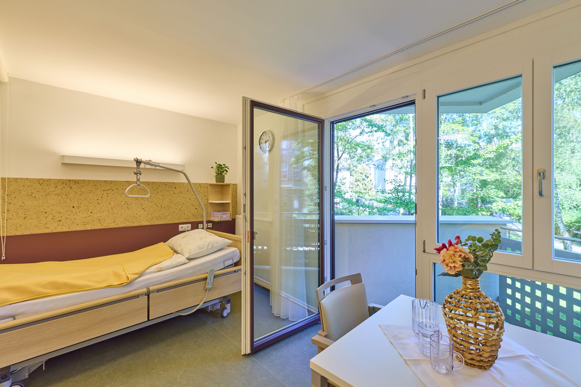Einzelzimmer in einer Pflegestation mit Pflegebett, Balkon und Tisch mit Sesseln.