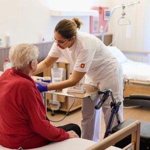 Eine Seniorin (von hinten, sitzend auf dem Bett) wird von einer Pflegerin betreut.