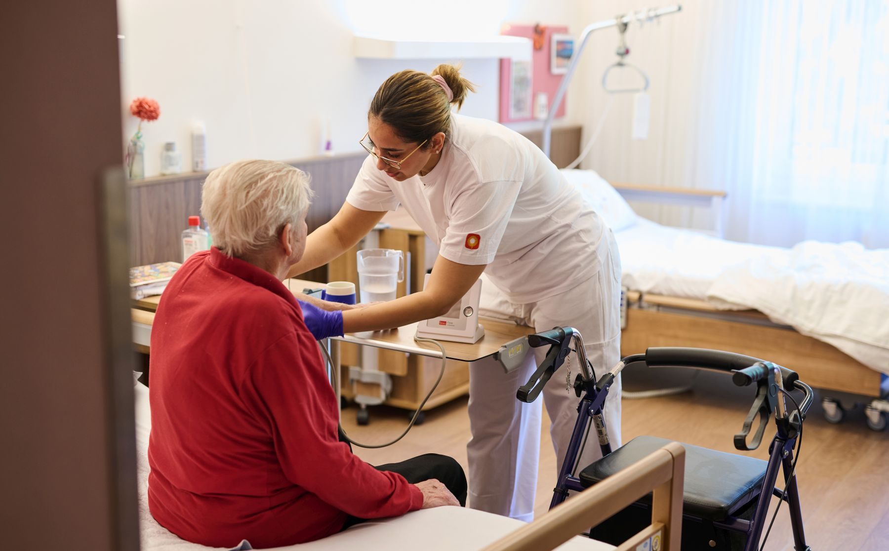 Eine Seniorin (von hinten, sitzend auf dem Bett) wird von einer Pflegerin betreut.