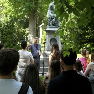 Ein Mann steht vor einem Grab am Zentralfriedhof und redet, Lehrlinge (von hinten) hören zu.