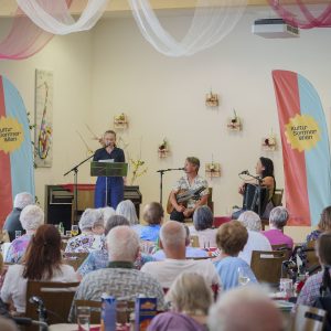Im Vordergrund Zuschauer*innen von hinten, im Hintergrund eine Bühne mit den Künstler*innen Gerti Drassl, Peter Havlicek und Marie-Theres Stickler beim Gartenkonzert im Rahmen von Kultursommer Wien im Haus Liebhartstal.