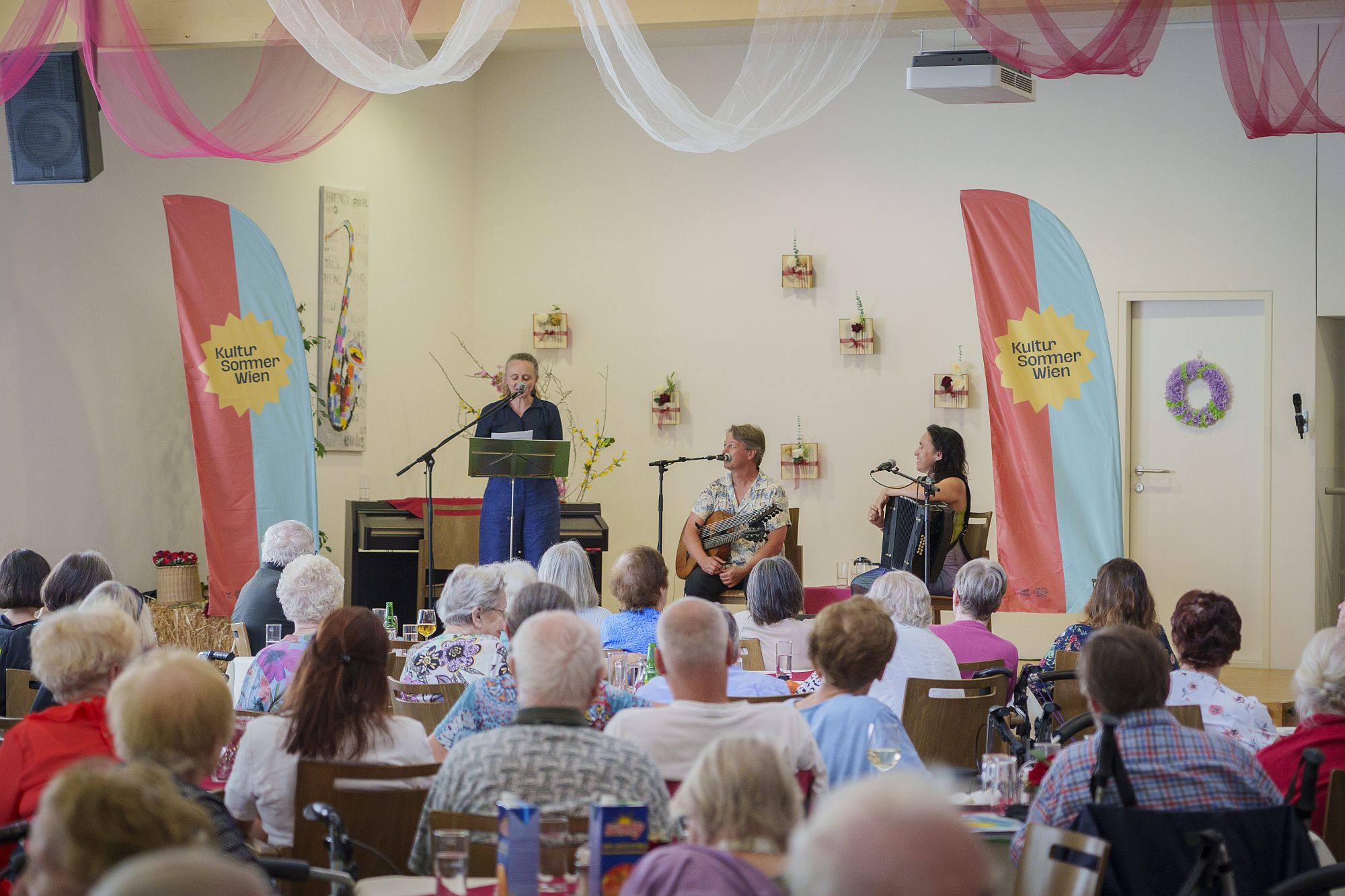 Im Vordergrund Zuschauer*innen von hinten, im Hintergrund eine Bühne mit den Künstler*innen Gerti Drassl, Peter Havlicek und Marie-Theres Stickler beim Gartenkonzert im Rahmen von Kultursommer Wien im Haus Liebhartstal.
