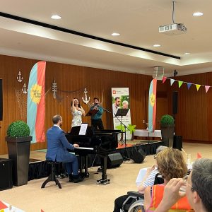 Eine Sängerin und ein Musiker auf einer Bühne und ein Klavierspieler vor der Bühne.