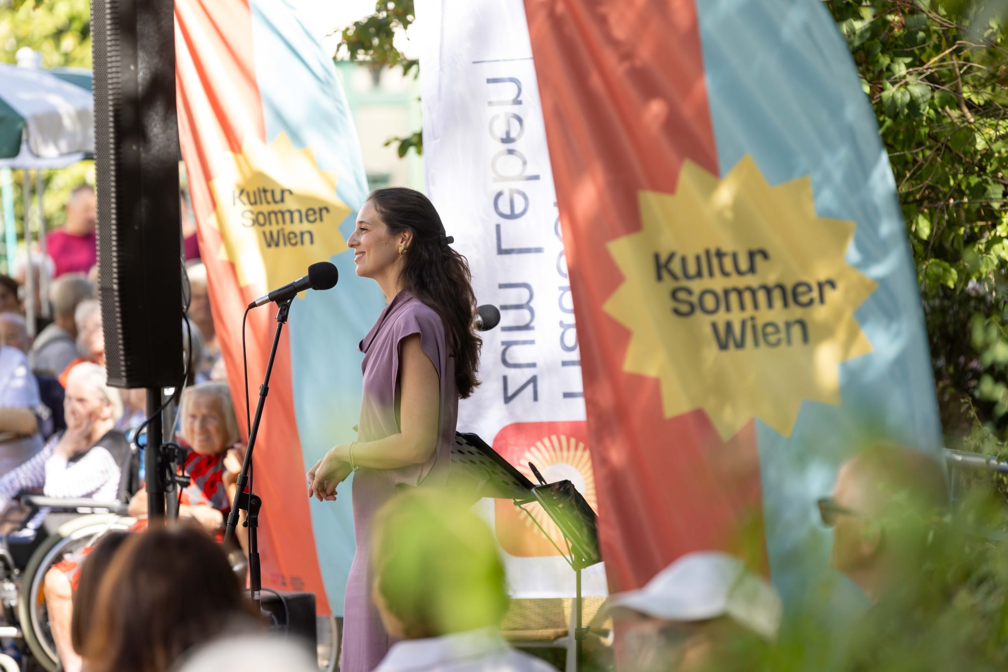 Ein Musikerin steht hinter einem Mikrofon auf der Bühne im Haus Hohe Warte. Im Hintergrund Beachflags mit der Aufschrift "Kultursommer" und "Häuser zum Leben"
