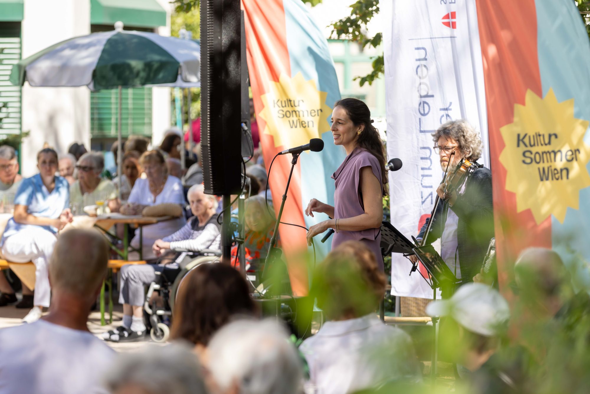 Eine Sängerin und ein Geiger auf einer Bühne im Garten des Hauses Hohe Warte beim Gartenkonzert im Rahmen von Kultursommer Wien.