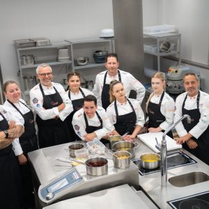 Zehn Köchinnen und Köche mit Kochschürzen in einer Großküche.