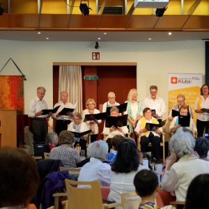 Ein Chor mit einer Klavierspielerin auf der Bühne, im Vordergrund Zuschauer*innen von hinten.
