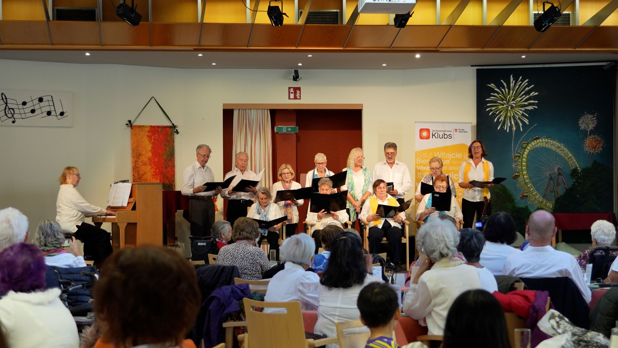 Ein Chor mit einer Klavierspielerin auf der Bühne, im Vordergrund Zuschauer*innen von hinten.