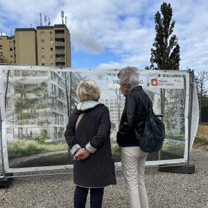 Zwei Frauen (von hinten) betrachten eine Plane, auf der ein Rendering des neuen Hauses Haidehof zu sehen ist.